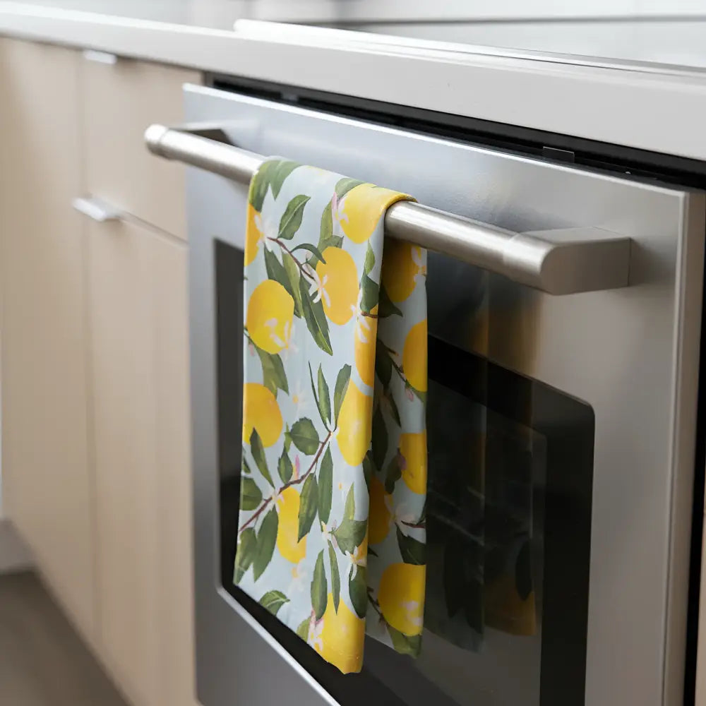 Artisan tea towel with lemon design features a vibrant yellow lemon and green leaf flour sack hanging on a stainless steel oven handle