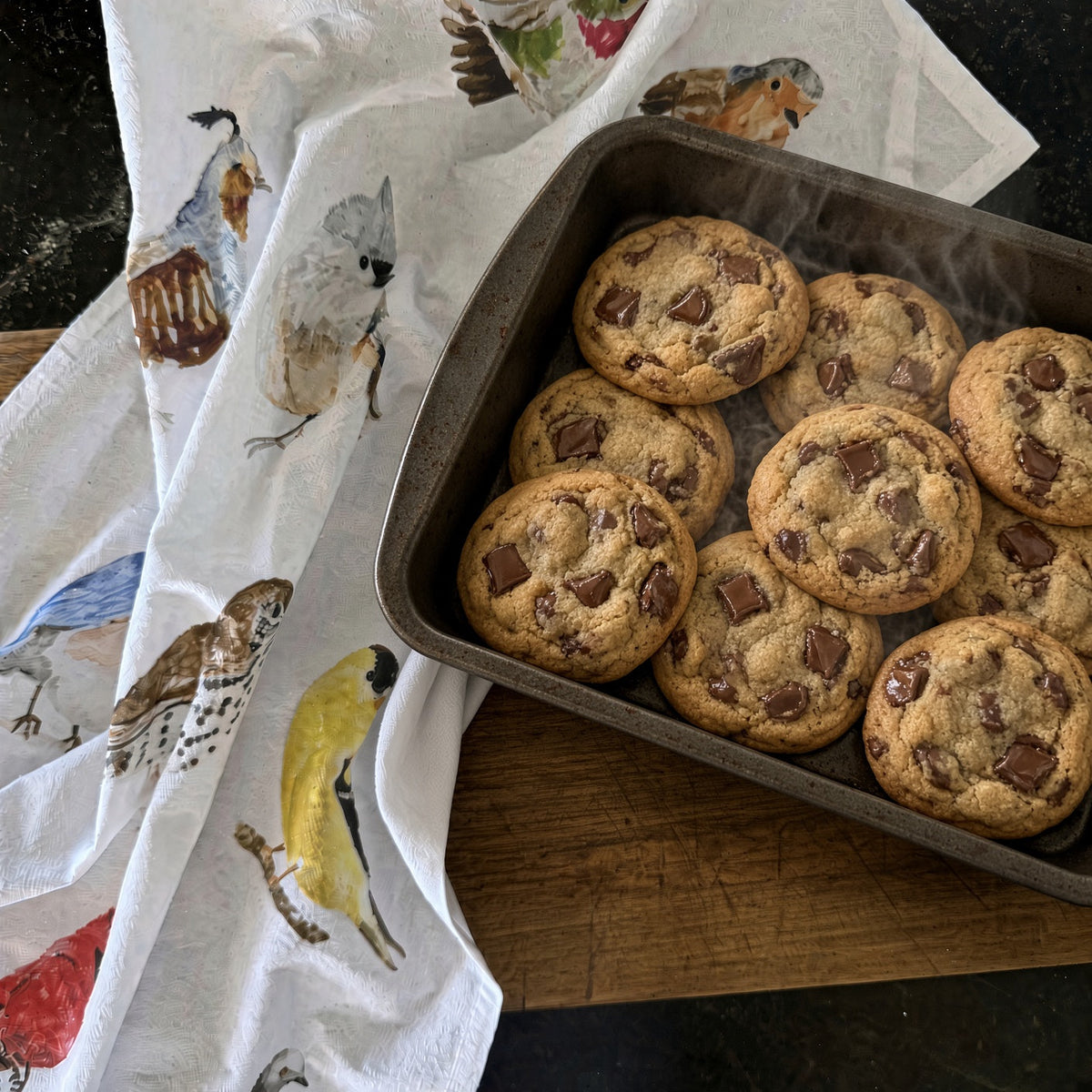 Golden-brown chocolate chip cookies in a dark metal pan on a birds cotton flour sack tea towel for baking use