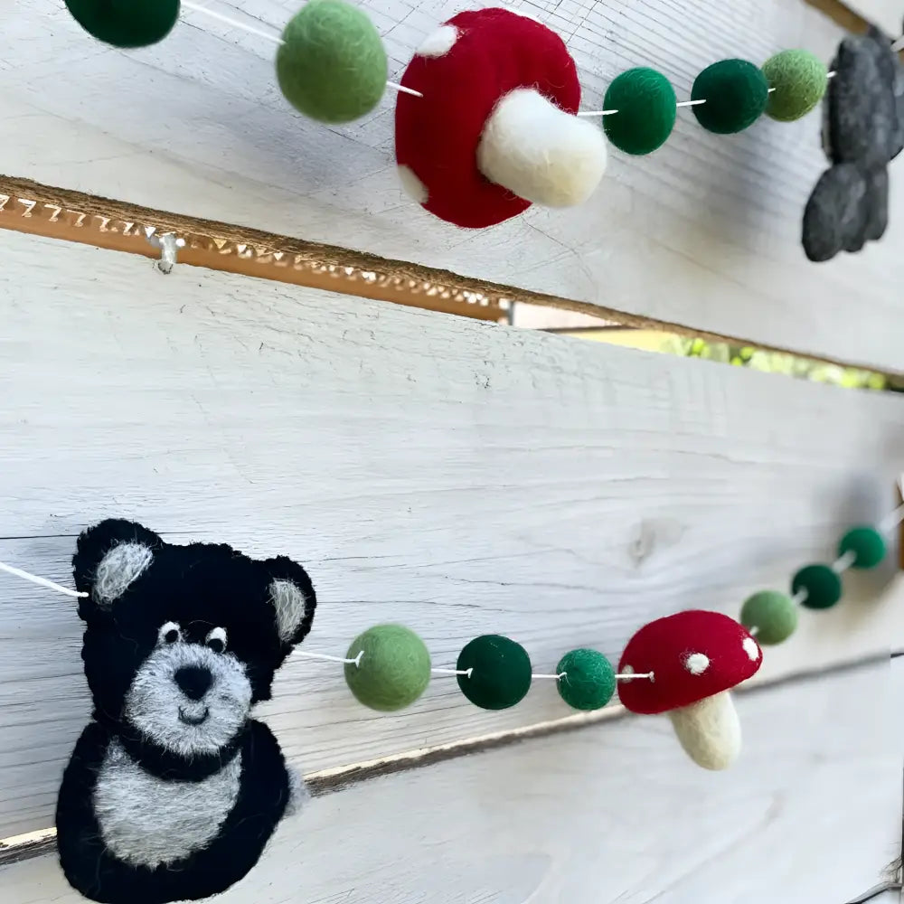 A hand-felted wool garland with whimsical forest friends featuring red green mushrooms black and gray bear faces and green balls