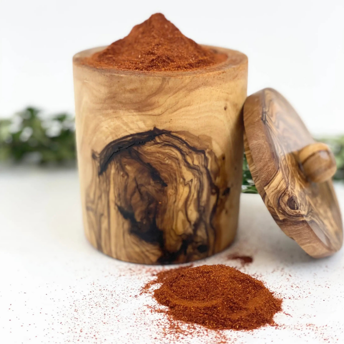 A cylindrical wooden spice jar filled with reddish-brown ground spice, shown with its lid leaning against the side on a white surface. A small pile of the spice is spilled in front, with blurred greenery in the background.