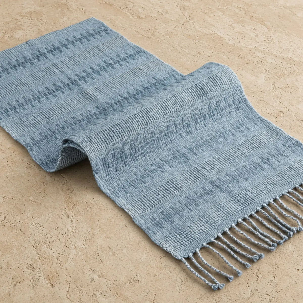 A soft light blue handwoven calado table runner with subtle geometric pattern and fringed edges made from recycled denim thread