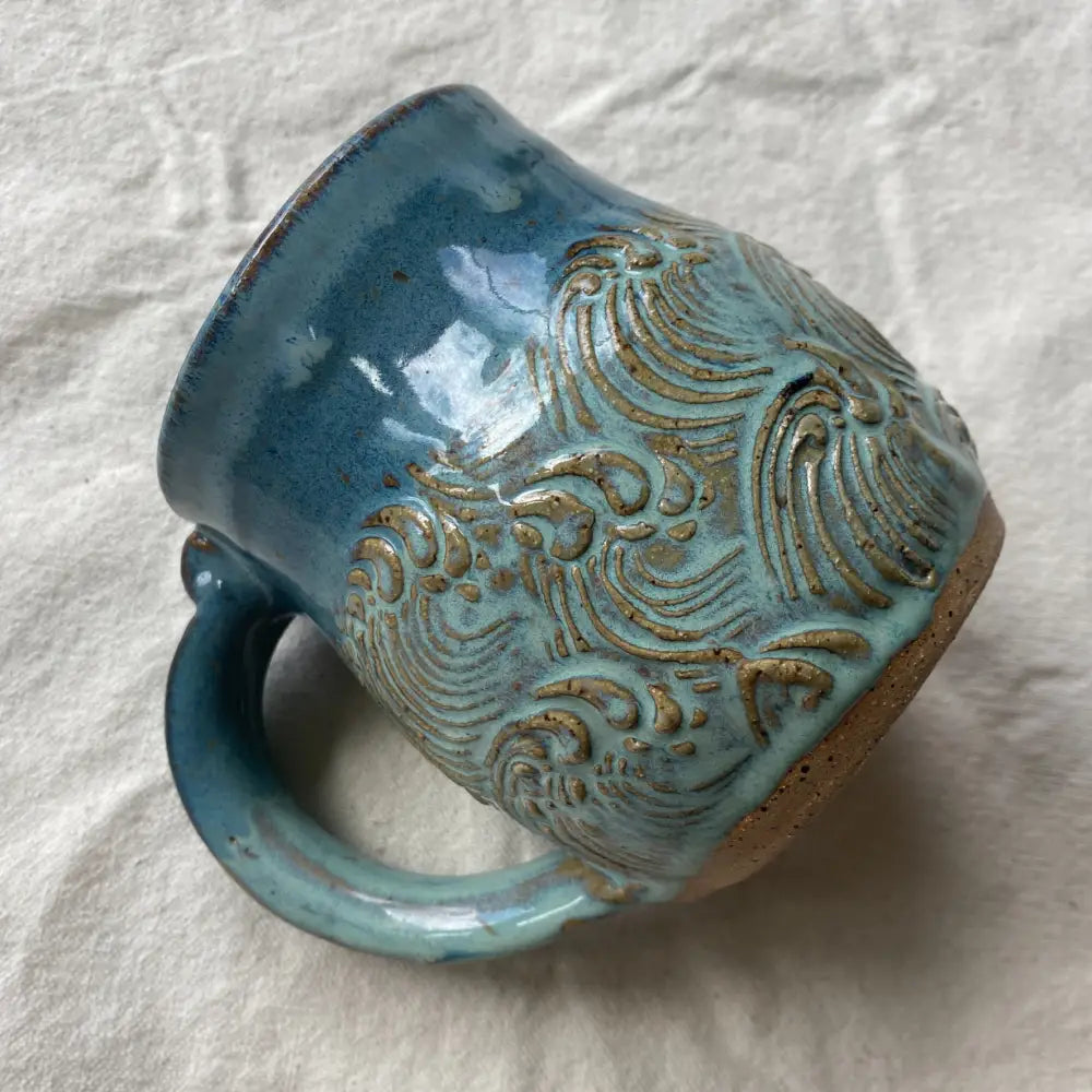 This handmade pottery mug features a textured sea-foam wave design with ocean blue rim.