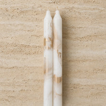 Two elegant white decorative taper candles with gold metallic swirls standing against a beige stone surface