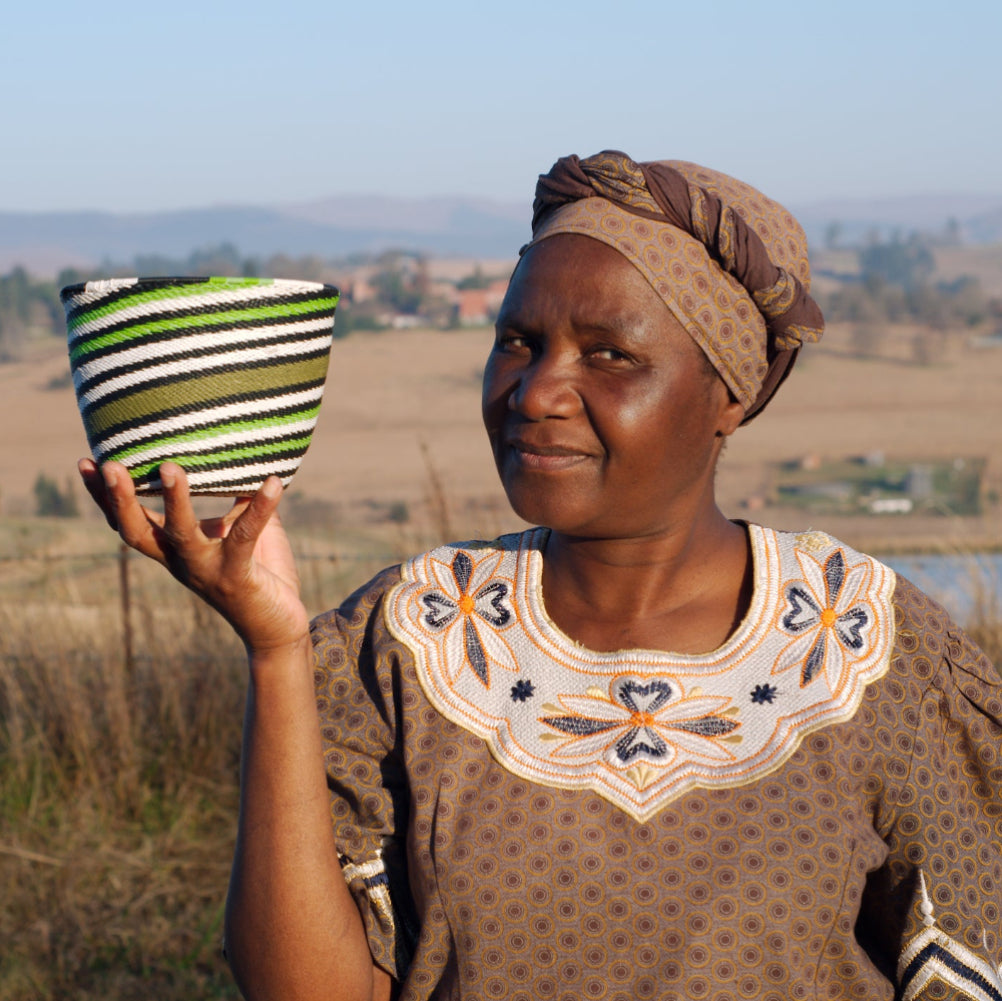 A woman outdoors wearing a brown patterned dress and headwrap, holding up a small woven basket with green, white, and black stripes.