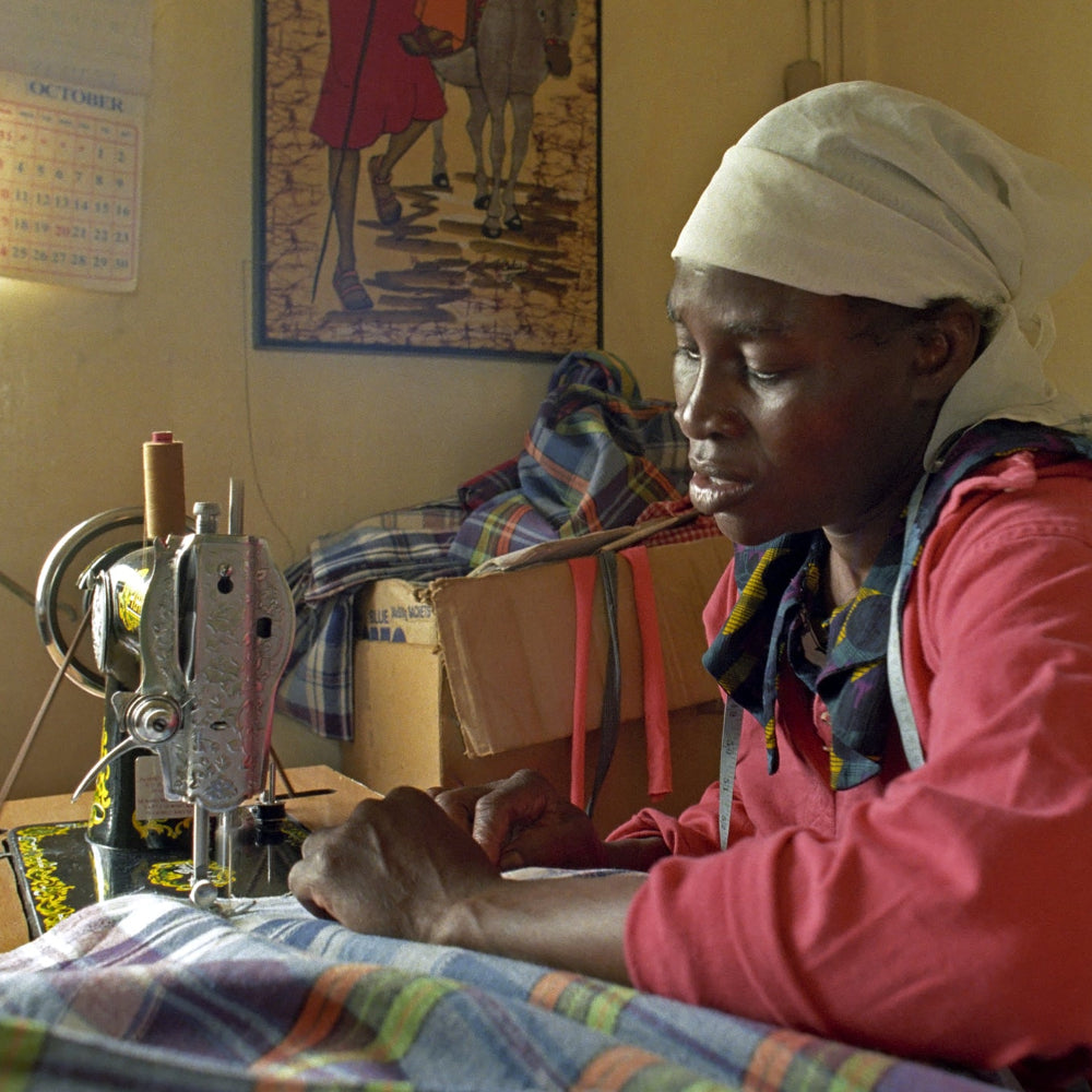 A woman wearing a white headscarf and a pink top sits at a sewing machine, stitching a plaid garment in a modest indoor workspace with walls, posters, and boxes visible.