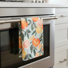 A soft light gray tea towel with a cheerful pattern of orange and yellow citrus fruits and green leaves