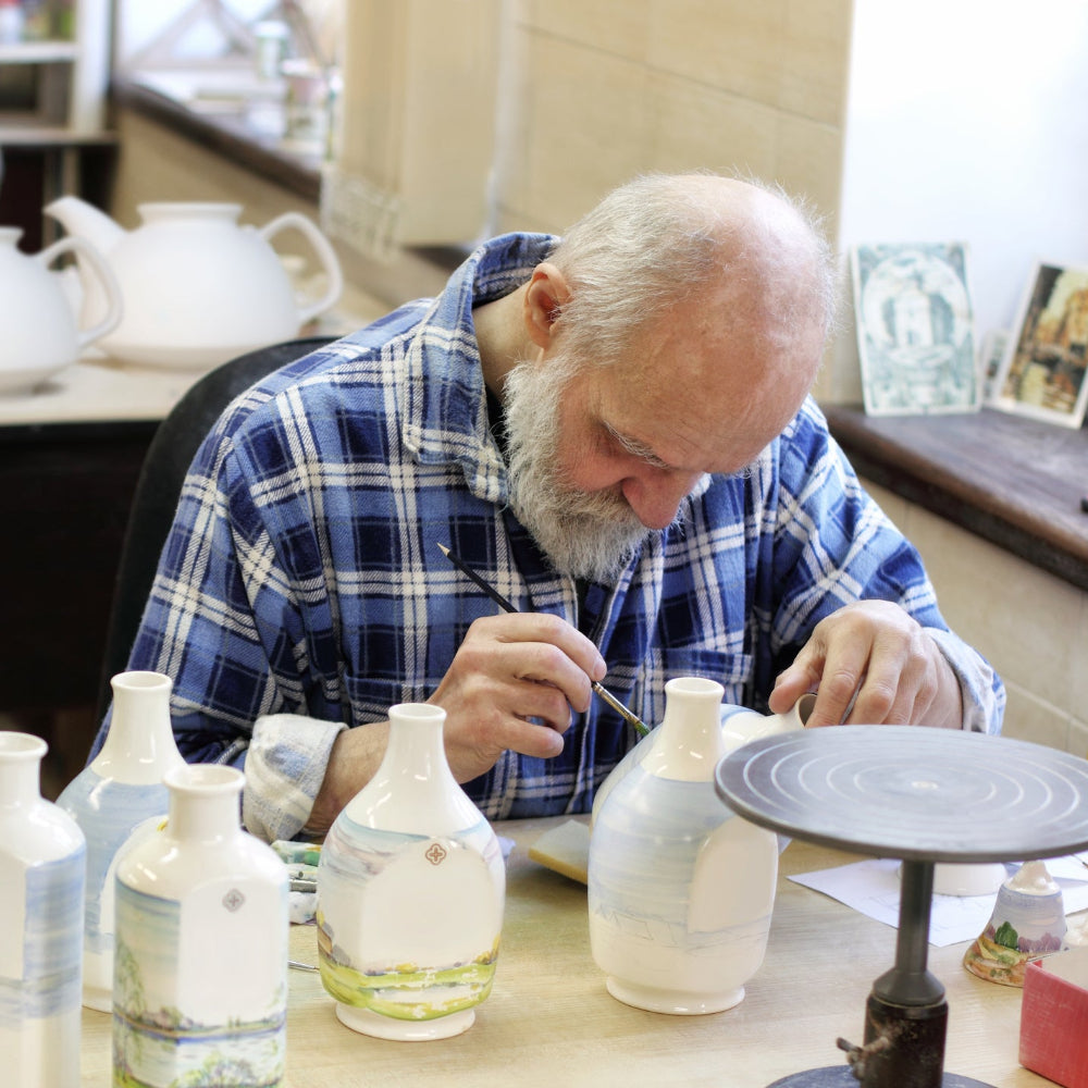 An elderly man with a white beard, wearing a blue and white plaid shirt, is seated at a pottery table, carefully painting ceramic vases. Several white and light blue vases with painted landscape designs are arranged on the table, with a pottery wheel and other ceramic pieces visible in the background workshop setting.