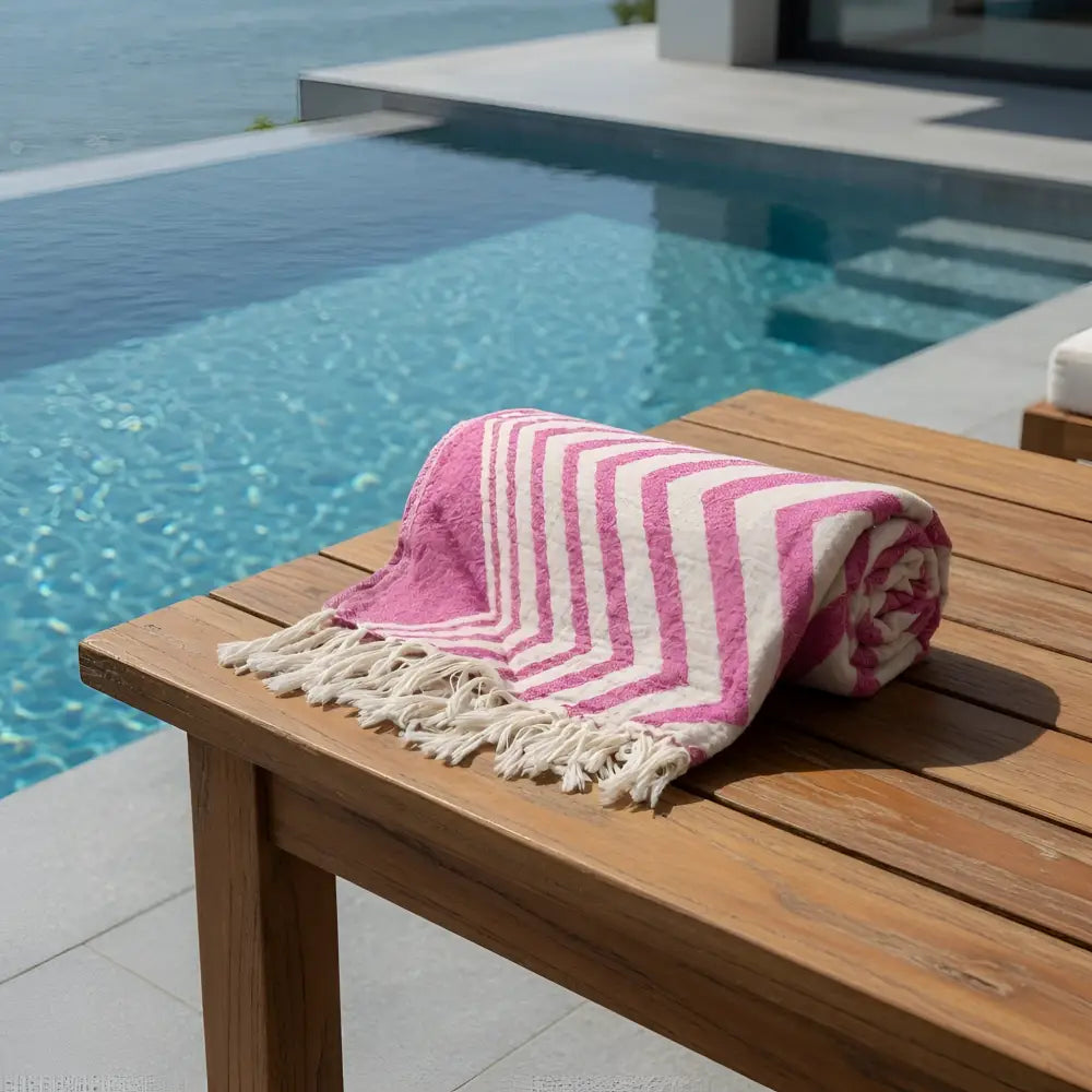 A rolled authentic turkish organic cotton beach towel in pink and white stripes with fringed edges on a wooden table by the pool