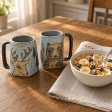 Two hand-painted stoneware mugs with deer and bear designs in rustic earthy tones and dark handles, sturdy and stylish
