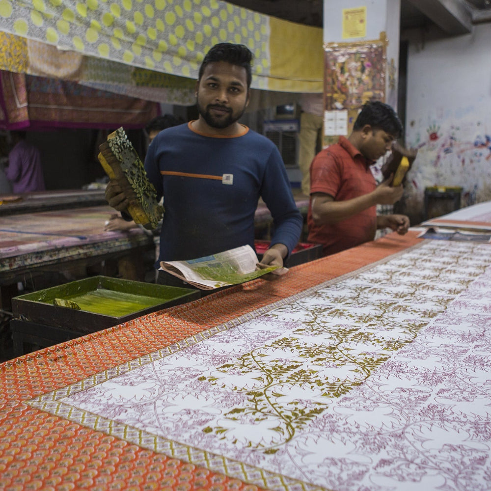 A large white and orange patterned fabric, likely a sari, is being printed with traditional motifs on a long table in a workshop. Two men are visible working behind the table, with other textile materials in the background.
