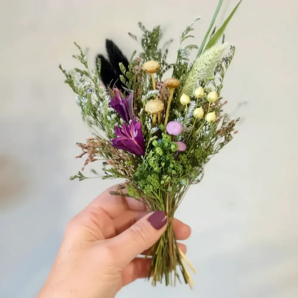 A hand holds a small dried floral bouquet with purple, yellow, and pink blooms tied with twine from bouquet in a box