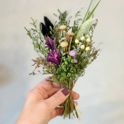A hand holds a small dried floral bouquet with purple, yellow, and pink blooms tied with twine from bouquet in a box