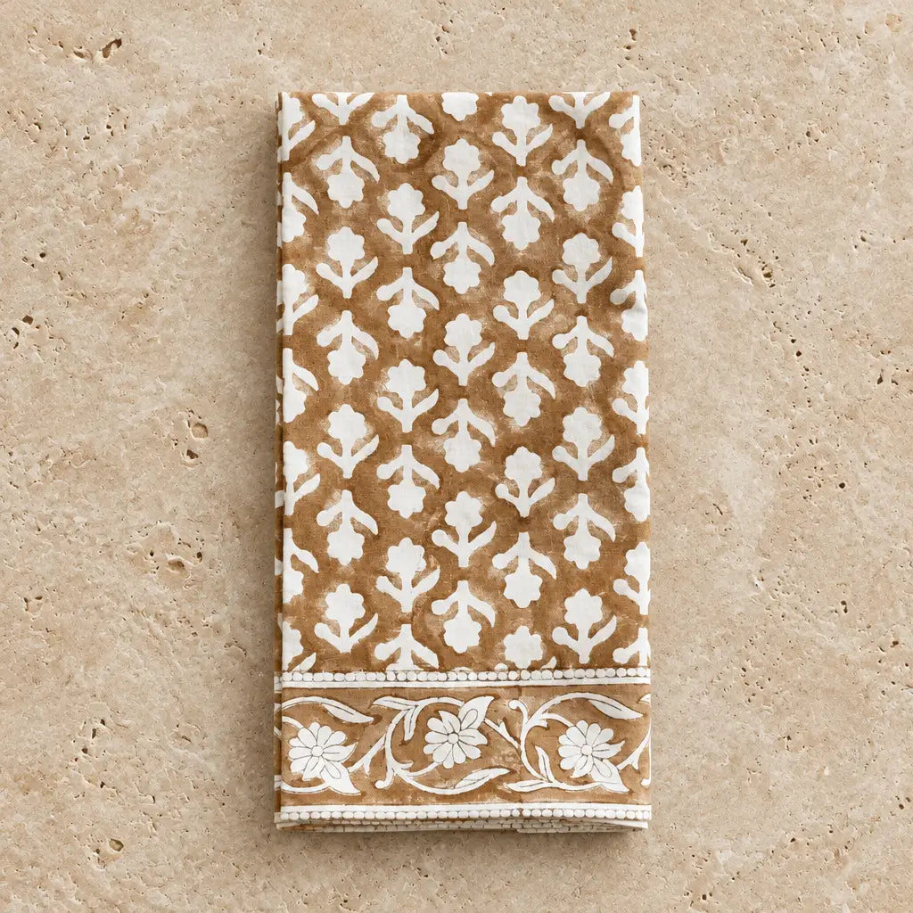 A folded caramel block print cotton napkin with brown and white floral patterned border in a set of 4