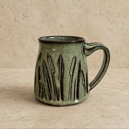 A handcrafted green ceramic handmade tapered mug with carved cattail pottery and textured vertical ridges