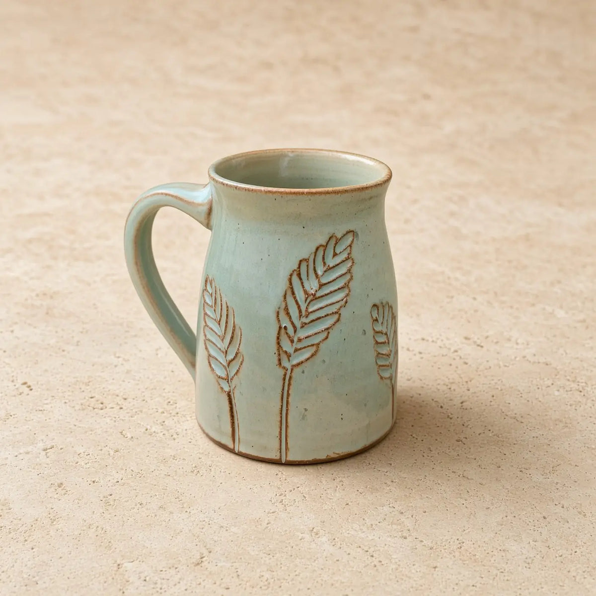 A light blue handmade tapered mug with a rustic finish and pottery carved leaf wheat stalks in warm brown tones