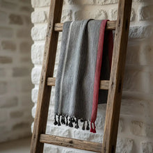 A soft charcoal white and ruby striped turkish cotton towel with black tassels hanging on a rustic wooden ladder