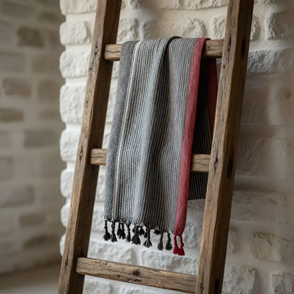 A soft charcoal white and ruby striped turkish cotton towel with black tassels hanging on a rustic wooden ladder