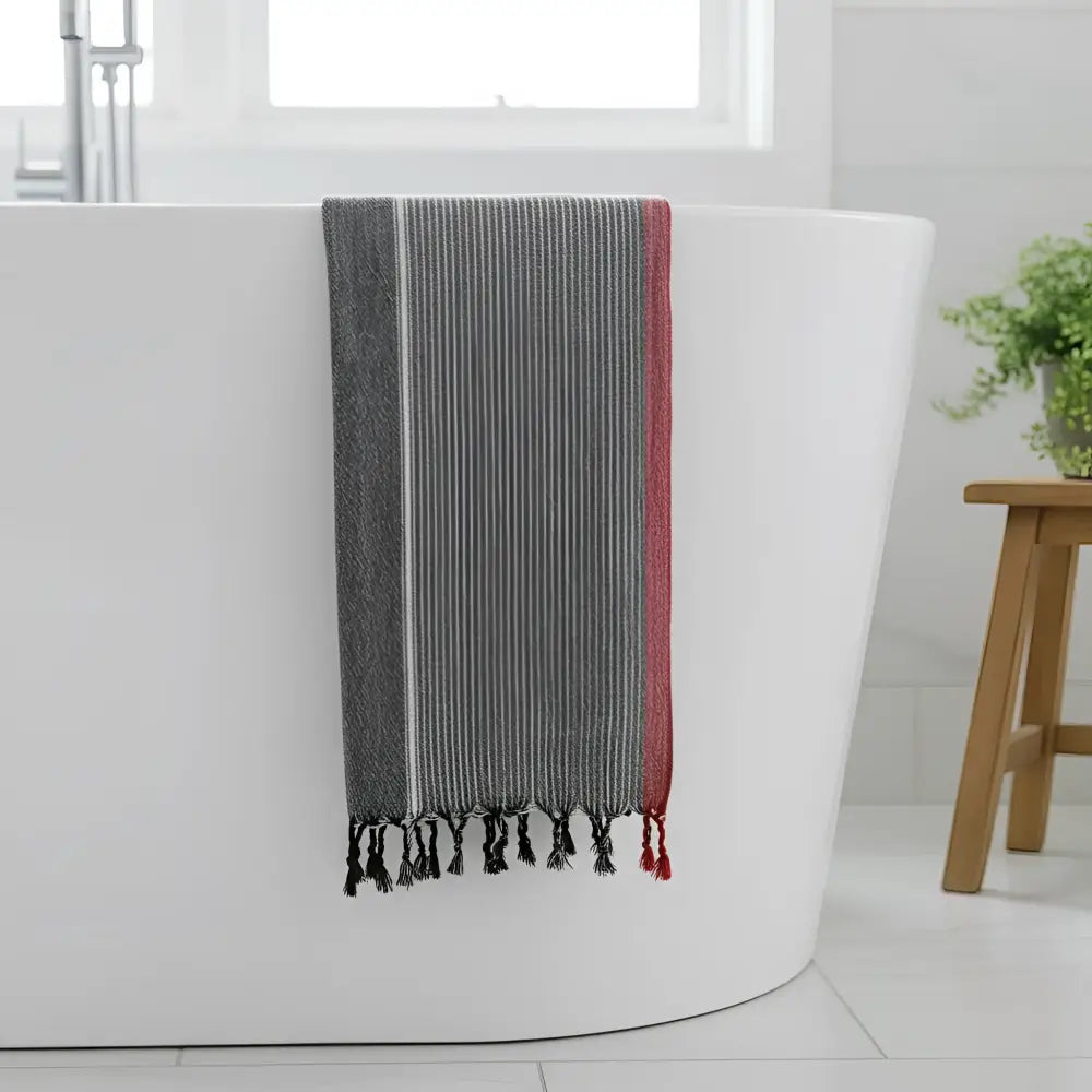 A charcoal white and ruby striped turkish hand towel with black tassels hanging over a white bathtub edge