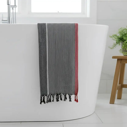 A charcoal white and ruby striped turkish hand towel with black tassels hanging over a white bathtub edge