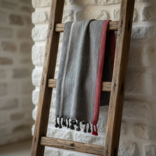 A soft charcoal white and ruby striped turkish cotton towel with black tassels hanging on a rustic wooden ladder