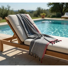 Charcoal white and ruby striped turkish cotton towel with red border and tassels draped on wooden lounge chair