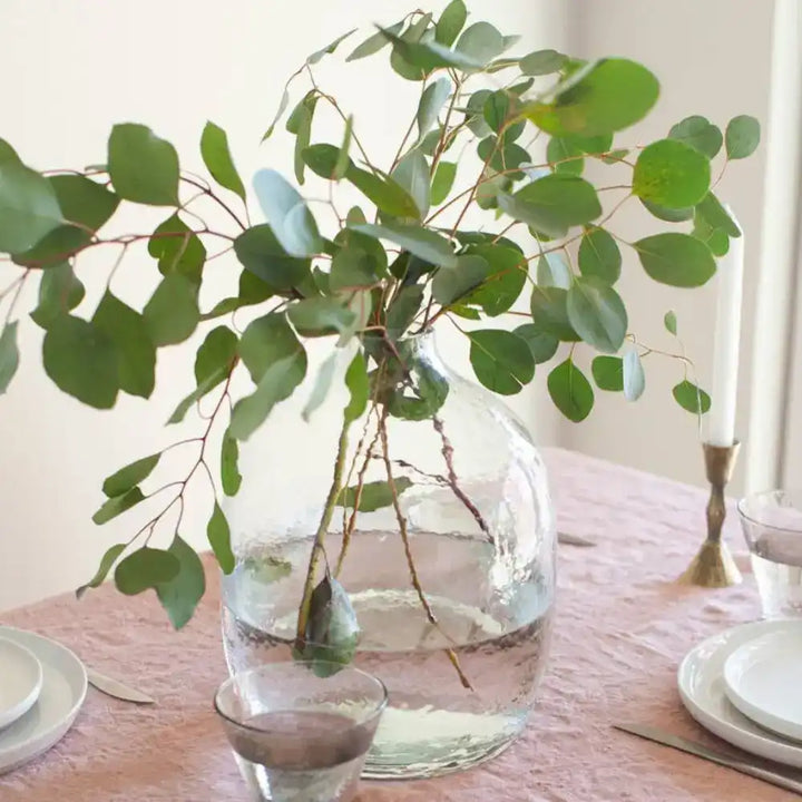 Handblown hammered glass vase in clear with sprigs of leaves on a table.
