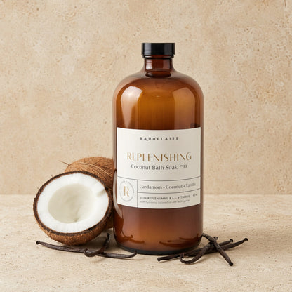 An amber glass bottle of baudelaire replenishing 40oz coconut bath soak with black cap beside coconut and vanilla beans for skin texture