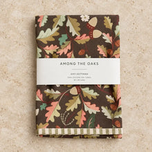 Folded brown cotton tea towel with whimsical leaf illustrations and cheerful striped edges from among the oaks collection