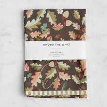 Folded brown cotton tea towel with whimsical leaf print of oak leaves acorns and flowers among the oaks