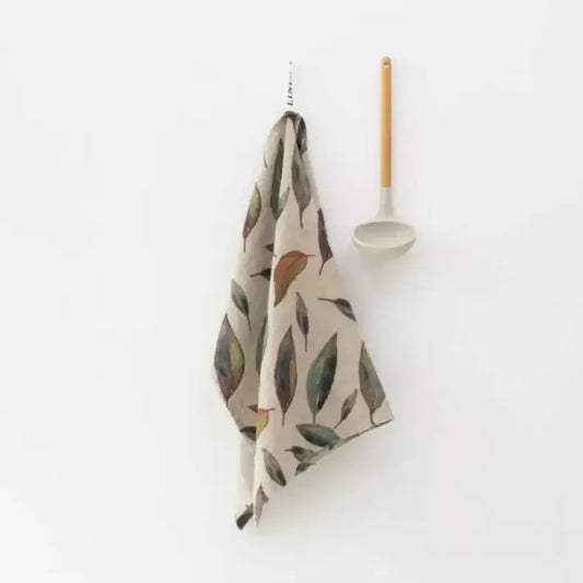 Kitchen towel with printed leaf design hanging against a white background next to a hanging ladle