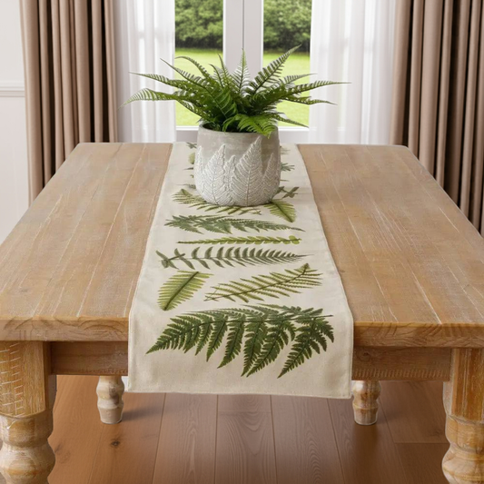 Table runner with fern design on a wooden table in a room with curtains and a plant.