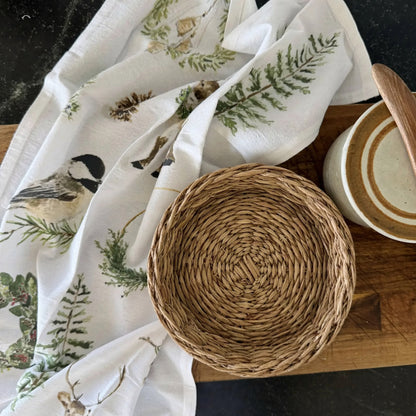 A round woven wicker basket in natural tan holding an evergreen cotton flour sack artisan tea towel