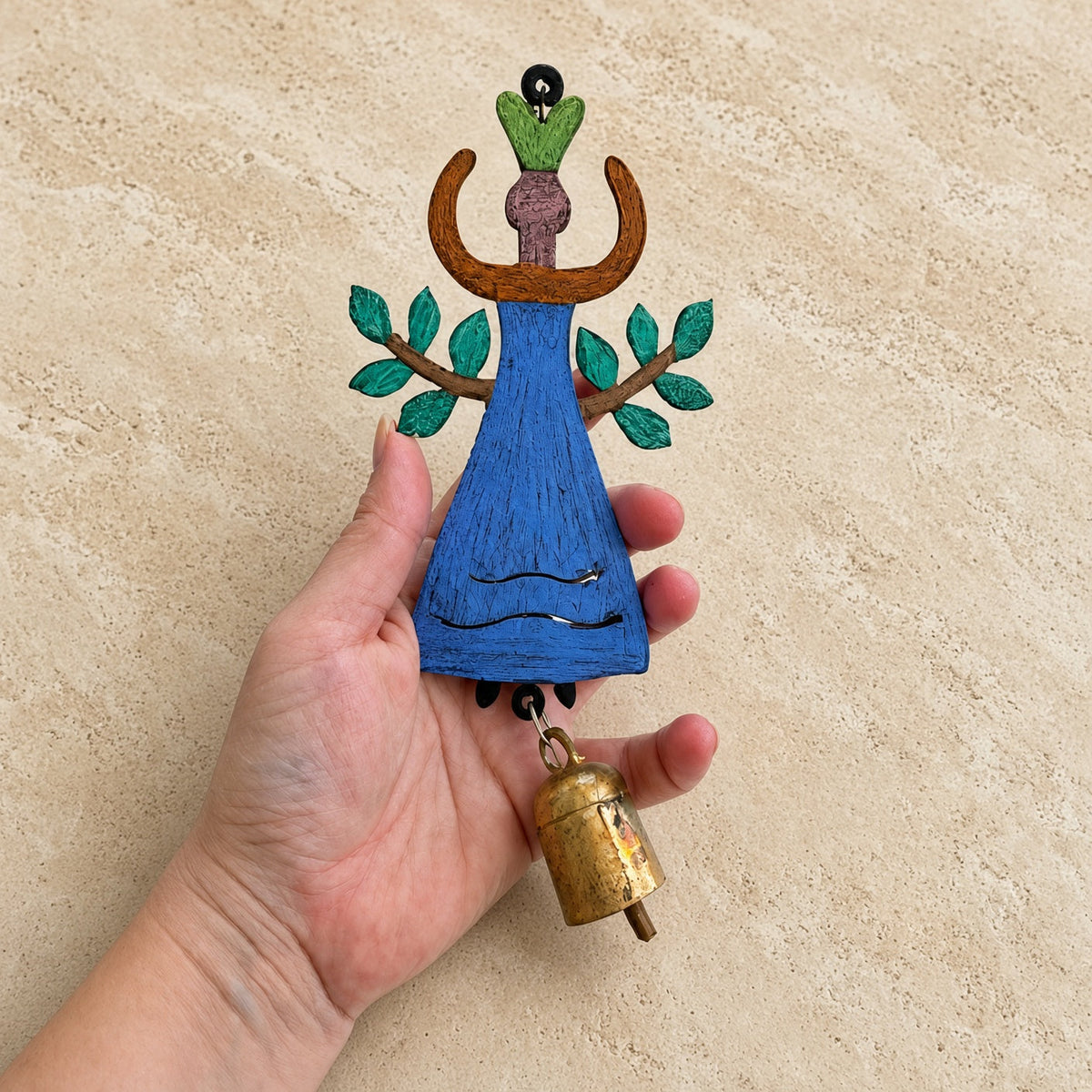 A hand holds a whimsical wooden garden angel bell chime ornament with blue figure green leafy arms and small brass bell