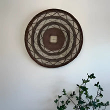Decorative woven wall art on a white wall with a plant in the corner.