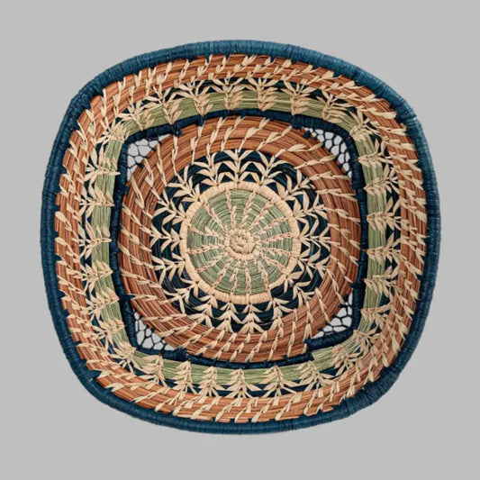 A basket featuring a variety of delicate raffia stitches on coils of pine needles and pajón, a native grass that grows in the highlands of Guatemala. A central circle transitions to a square basket. Indigo blue raffia and pine needles accent the lovely design