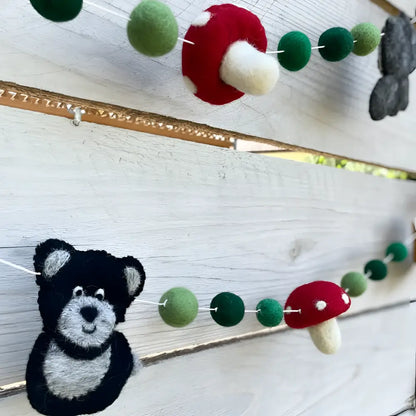 A hand-felted wool garland with whimsical forest friends featuring red green mushrooms black and gray bear faces and green balls