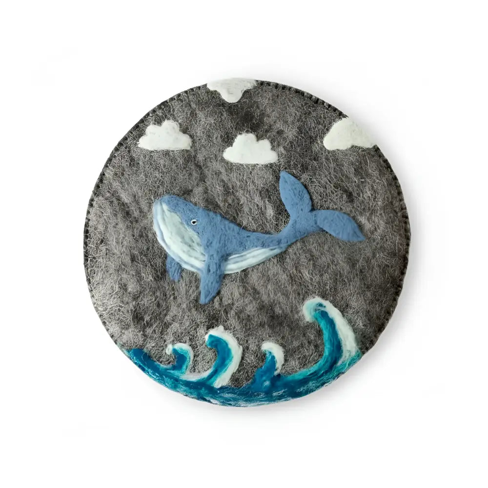 Gray hand-felted wool trivet featuring a blue whale swimming among stylized waves and clouds with needle-felted details