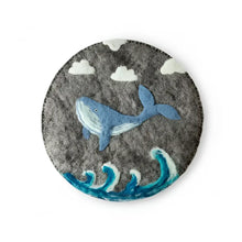Gray hand-felted wool trivet featuring a blue whale swimming among stylized waves and clouds with needle-felted details