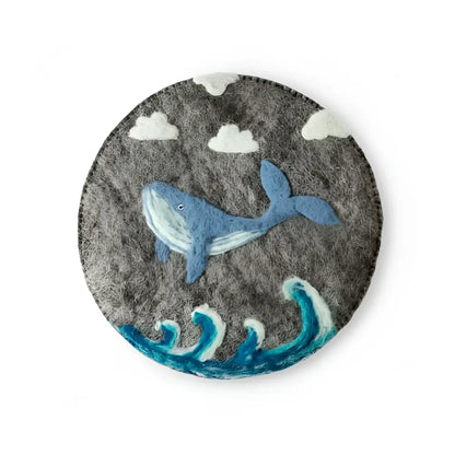 Gray hand-felted wool trivet featuring a blue whale swimming among stylized waves and clouds with needle-felted details