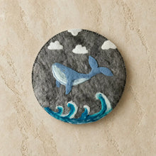 Circular hand-felted wool trivet with a charming blue whale above stylized ocean waves in soft greys and blue