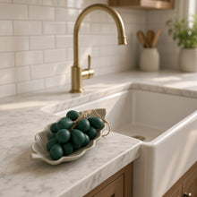 A white ceramic leaf bowl holding dark green twine tied decorative eggs next to handcrafted grape cluster olive oil soap