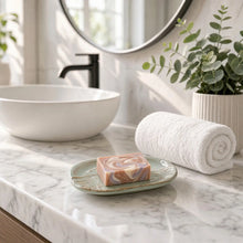 A beautiful handmade soap with pink and lavender swirls sits on a handcrafted stoneware dish with lace pattern