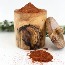 A cylindrical wooden spice jar filled with reddish-brown ground spice, shown with its lid leaning against the side on a white surface. A small pile of the spice is spilled in front, with blurred greenery in the background.