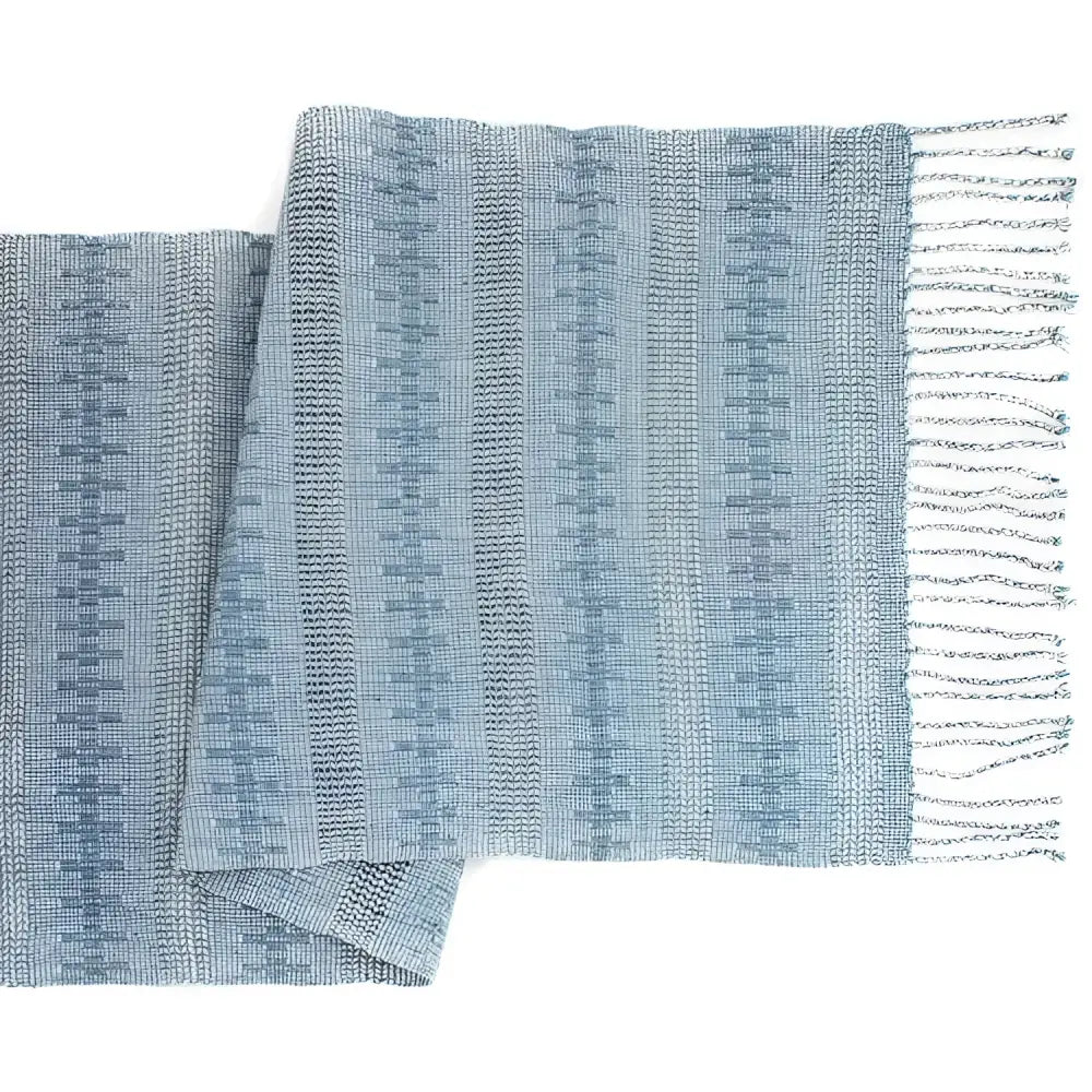 Unique table runner is handwoven and finished with hand-twisted fringe. It has subtle blue recycled denim thread paired with the airy open weave technique known as calado.
