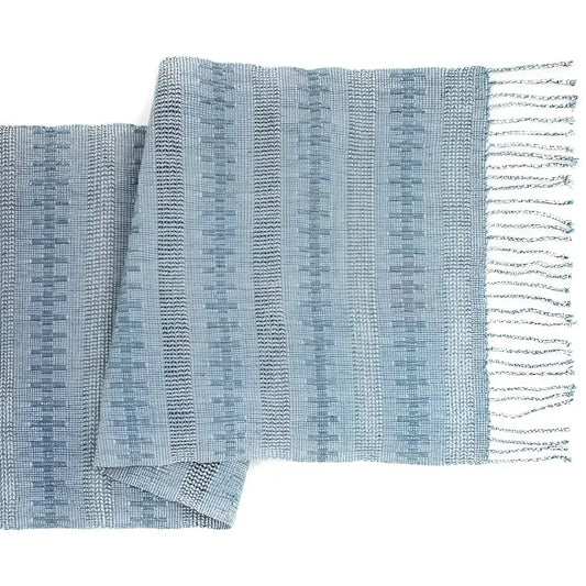 Unique table runner is handwoven and finished with hand-twisted fringe. It has subtle blue recycled denim thread paired with the airy open weave technique known as calado.