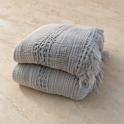 Stacked light gray handwoven waffle bed blankets with fringed edges made of cotton for cozy use