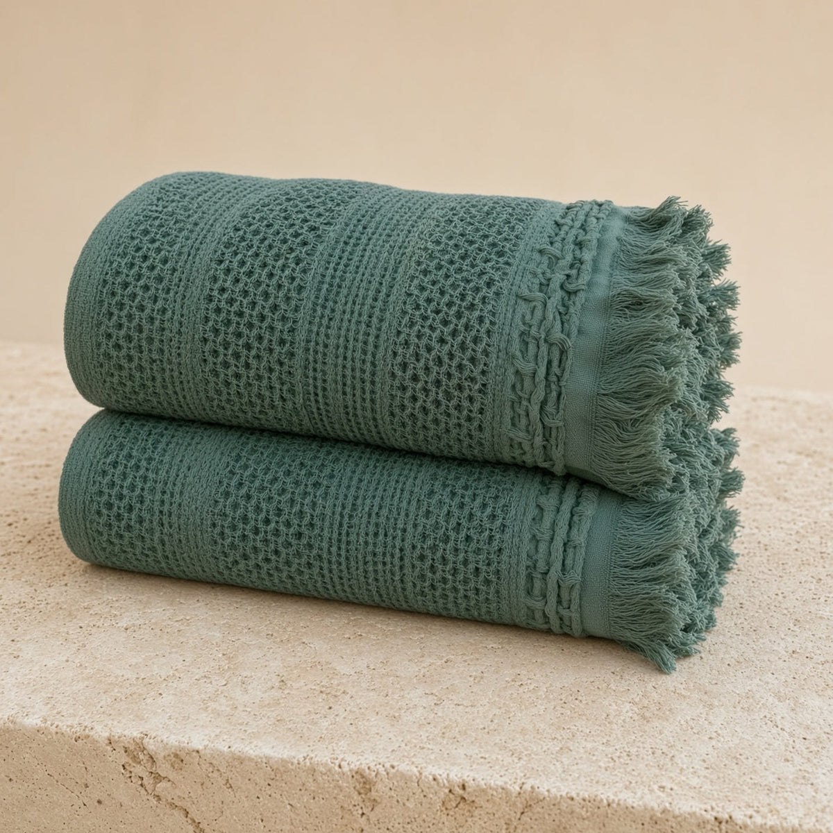 Two rolled emerald green handwoven waffle bed cotton towels with fringed edges and braided trim next to waffle bed blanket