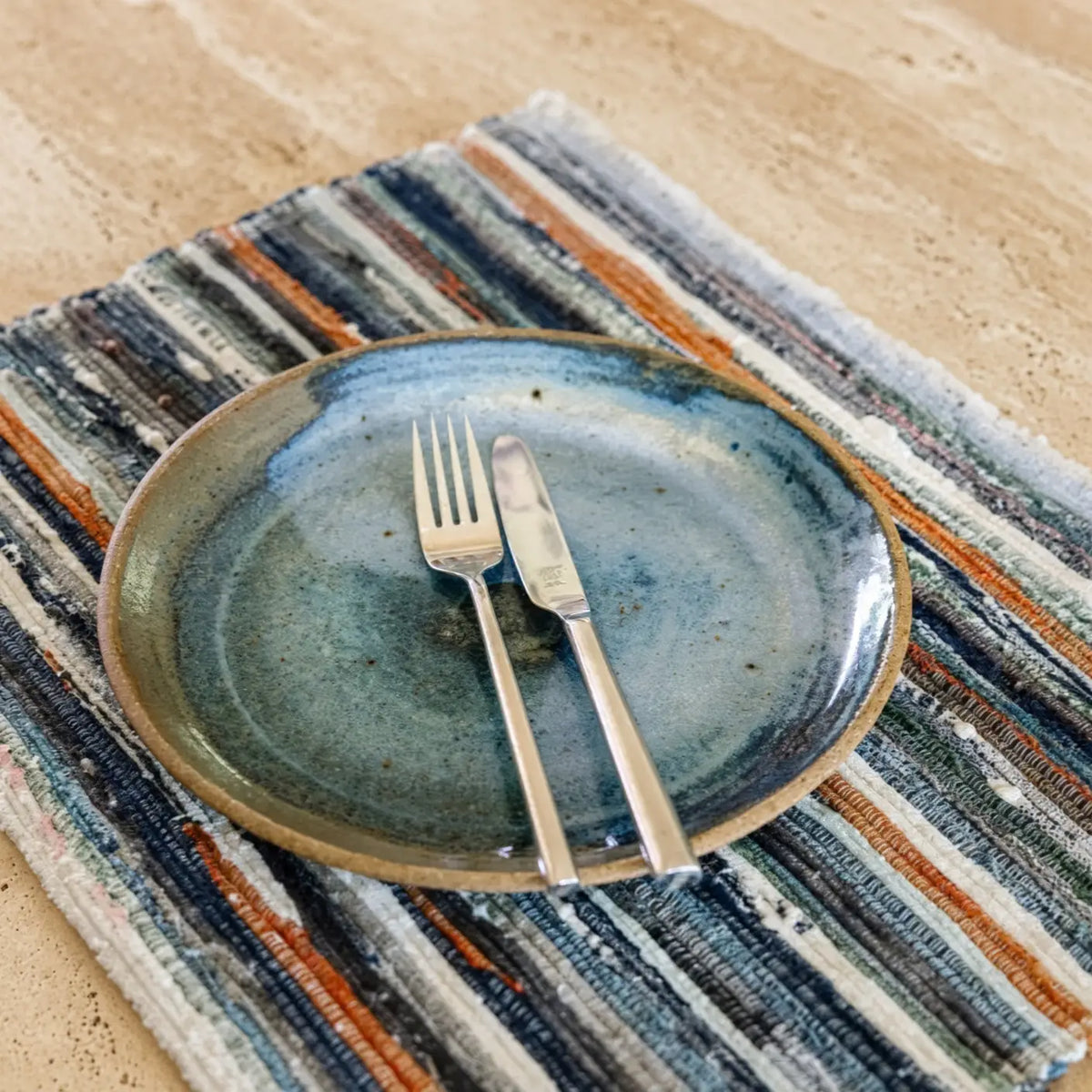 Handcrafted ceramic plate with mottled blue glaze and earthy rim on handwoven upcycled fabric placemat with stripes