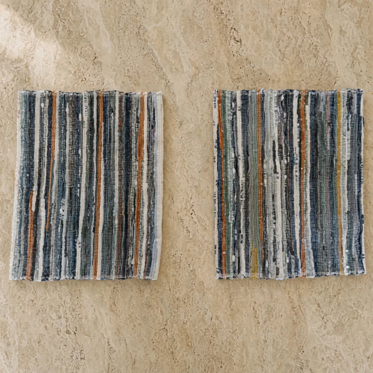 Two handwoven upcycled fabric placemats made from recycled denim strips in navy blue gray white and earthy orange tones