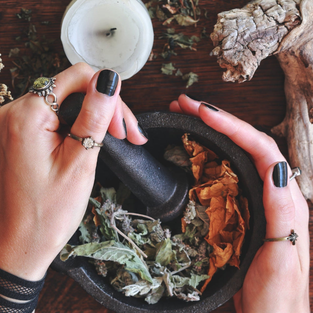 A top-down view of a person's hands with black nail polish holding a black mortar and pestle filled with dried herbs and leaves, surrounded by candles, dried flowers, and natural wood on a dark wooden table.